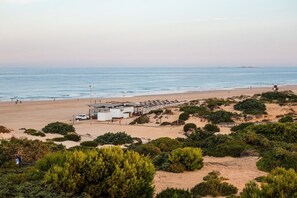 A beach with a lifeguard tower, a white building with a flat roof, and dense greenery in the foreground. - GuestReady - Chiclana Labarrosa (Chiclana de la Frontera)