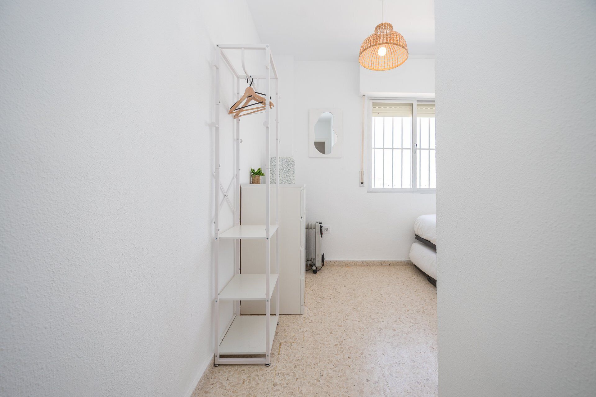 Een kleine kamer met witte wanden, een bed, een radiator, een hanglamp en een raam met blinds.