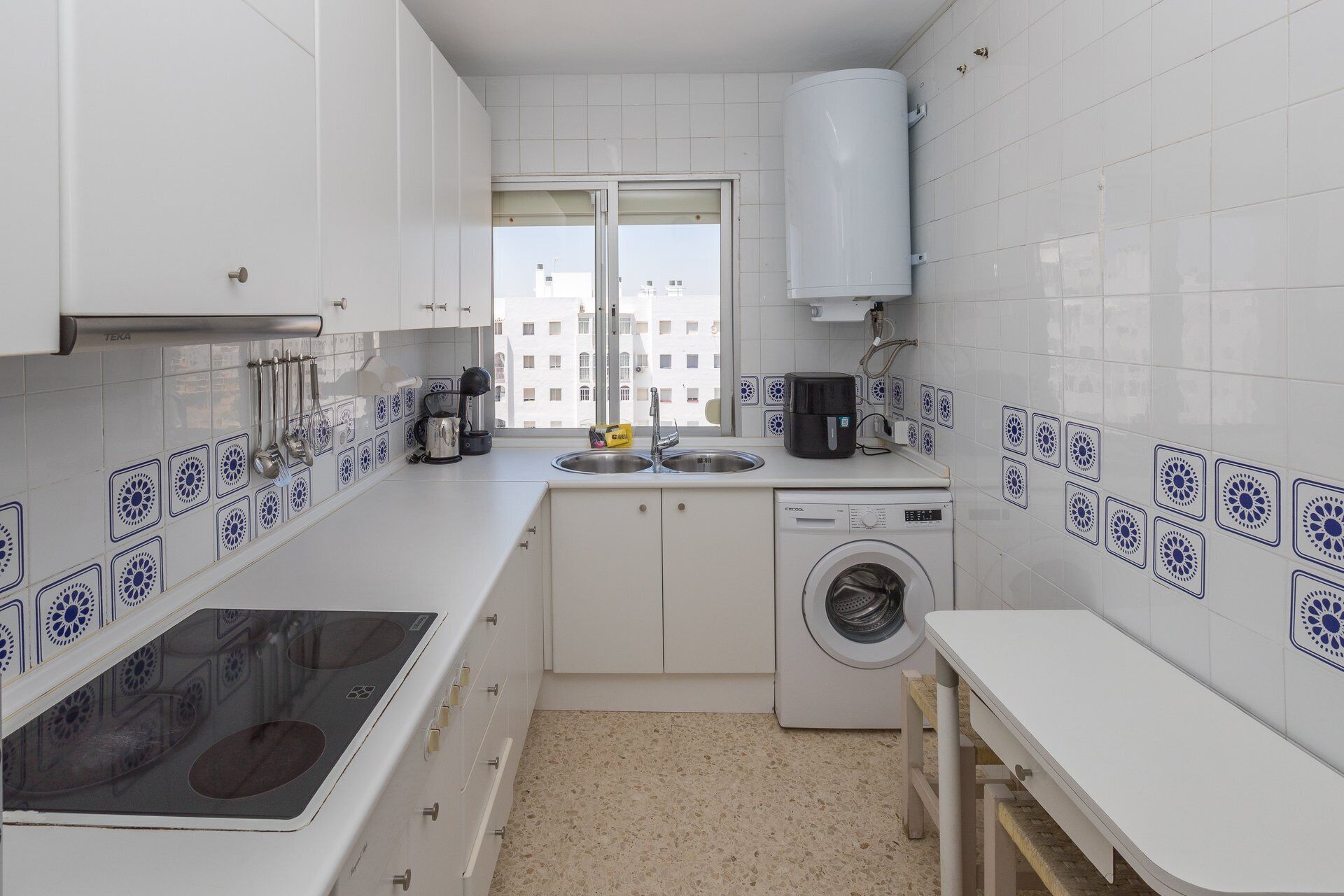 Una cocina compacta con muebles blancos, lavadora integrada y ventana con vista a edificios.