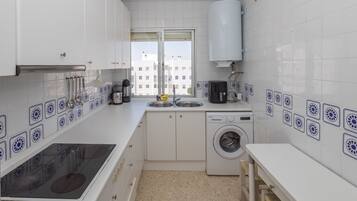 Una cocina compacta con muebles blancos, lavadora integrada y ventana con vista a edificios.