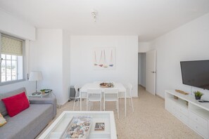 Ein modernes Wohnzimmer mit einem weißen Sofa, einem Esstisch mit Stühlen, einem Fernseher auf einem Standfuß und einem Bild an der Wand.