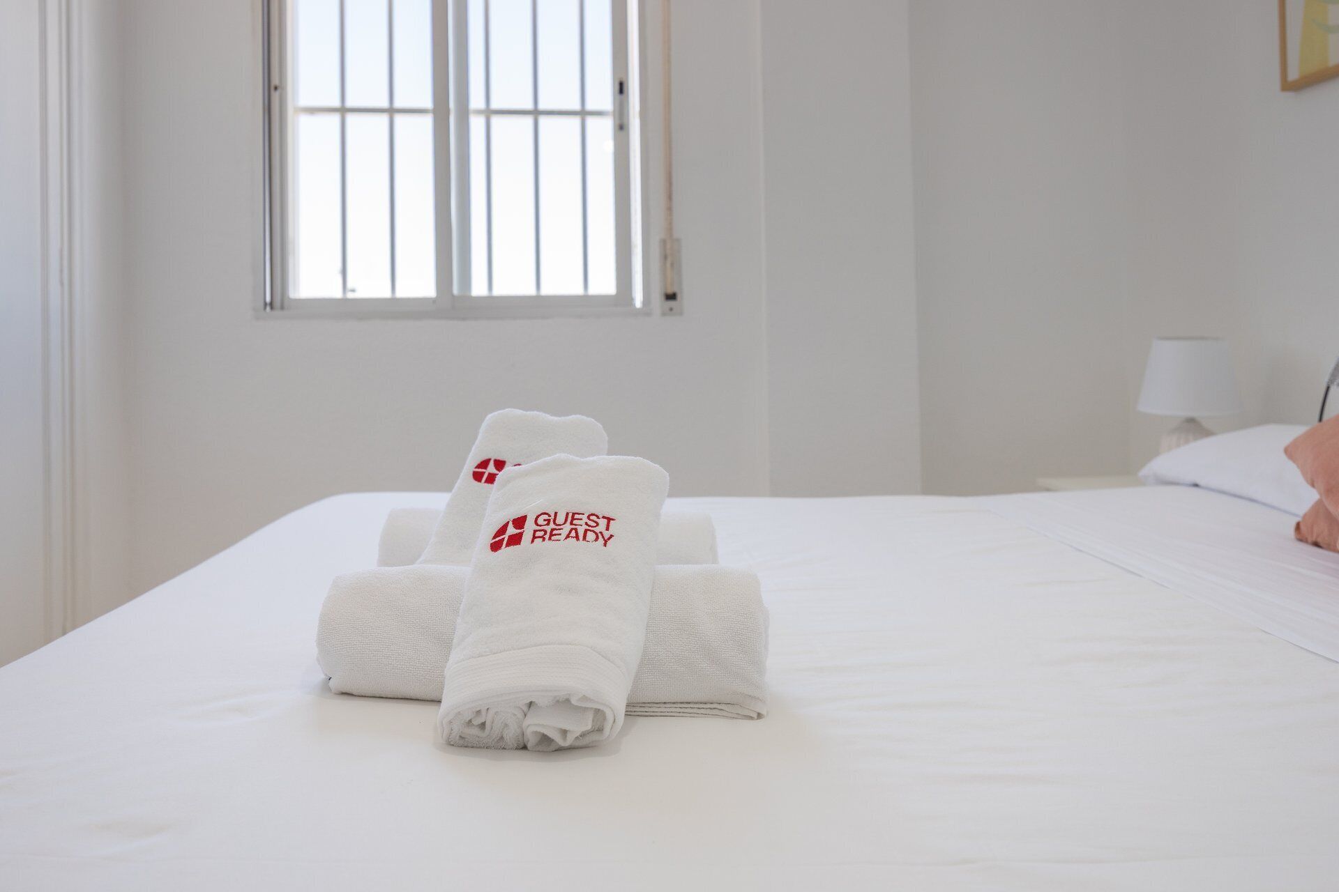 A neatly made bed with a folded towel in the center and a window with blinds in the background.