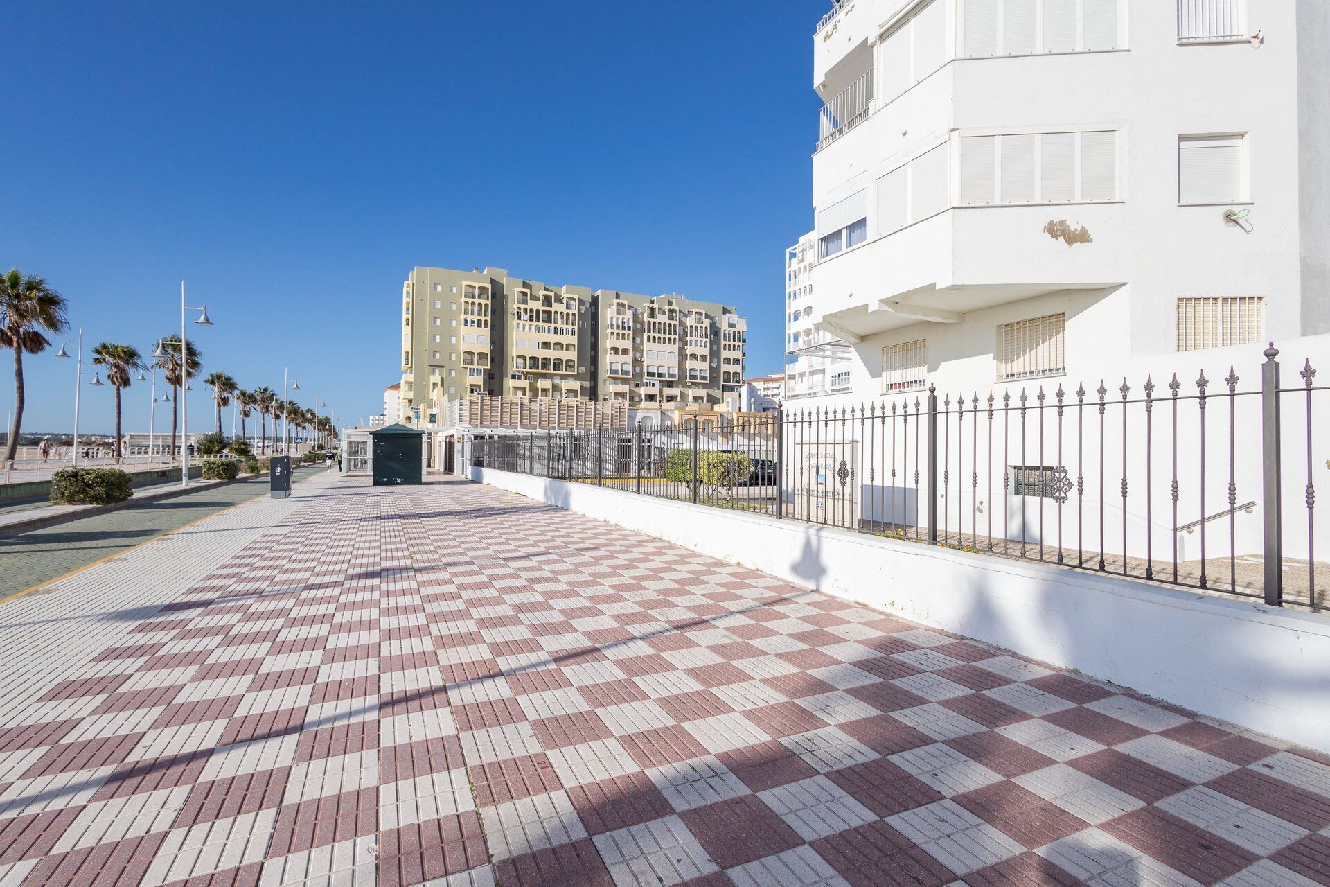 A paved walkway with a checkered pattern, bordered by a white building and a metal fence.