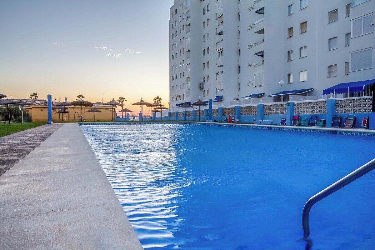 A swimming pool with a clear blue water surface, surrounded by a concrete deck and a tall building in the background.