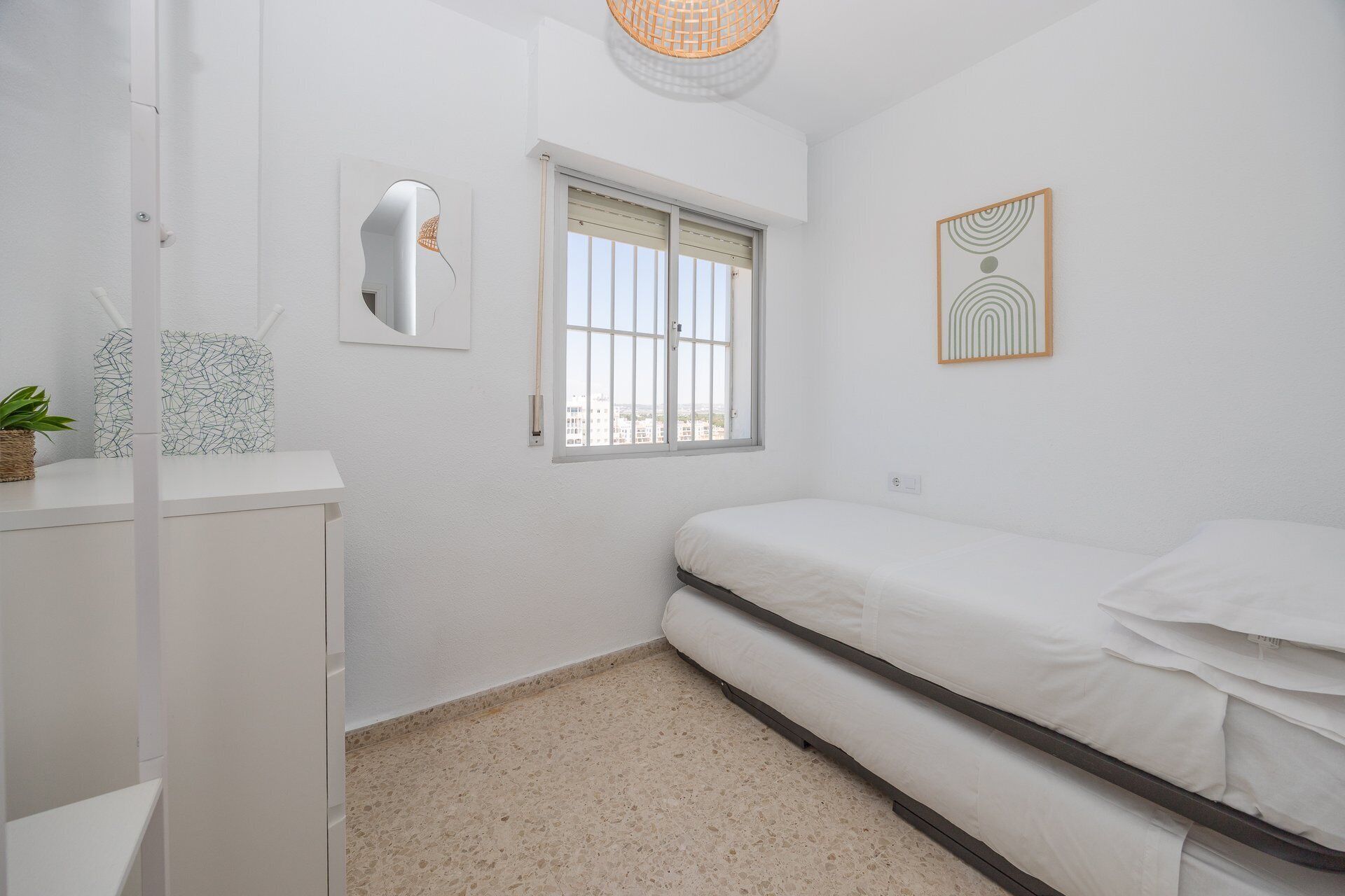 Ein Schlafzimmer mit einem Bett, einem Fenster, einem an der Wand befestigten Spiegel und einer kleinen Pflanze auf einem Regal.