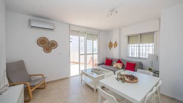 Ein modernes Wohnzimmer mit einer Couch, einem Esstisch und einem Sessel. Es gibt ein Fenster mit Jalousien, einen Deckenventilator und eine Klimaanlage.