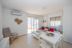 Ein modernes Wohnzimmer mit einer Couch, einem Esstisch und einem Sessel. Es gibt ein Fenster mit Jalousien, einen Deckenventilator und eine Klimaanlage.
