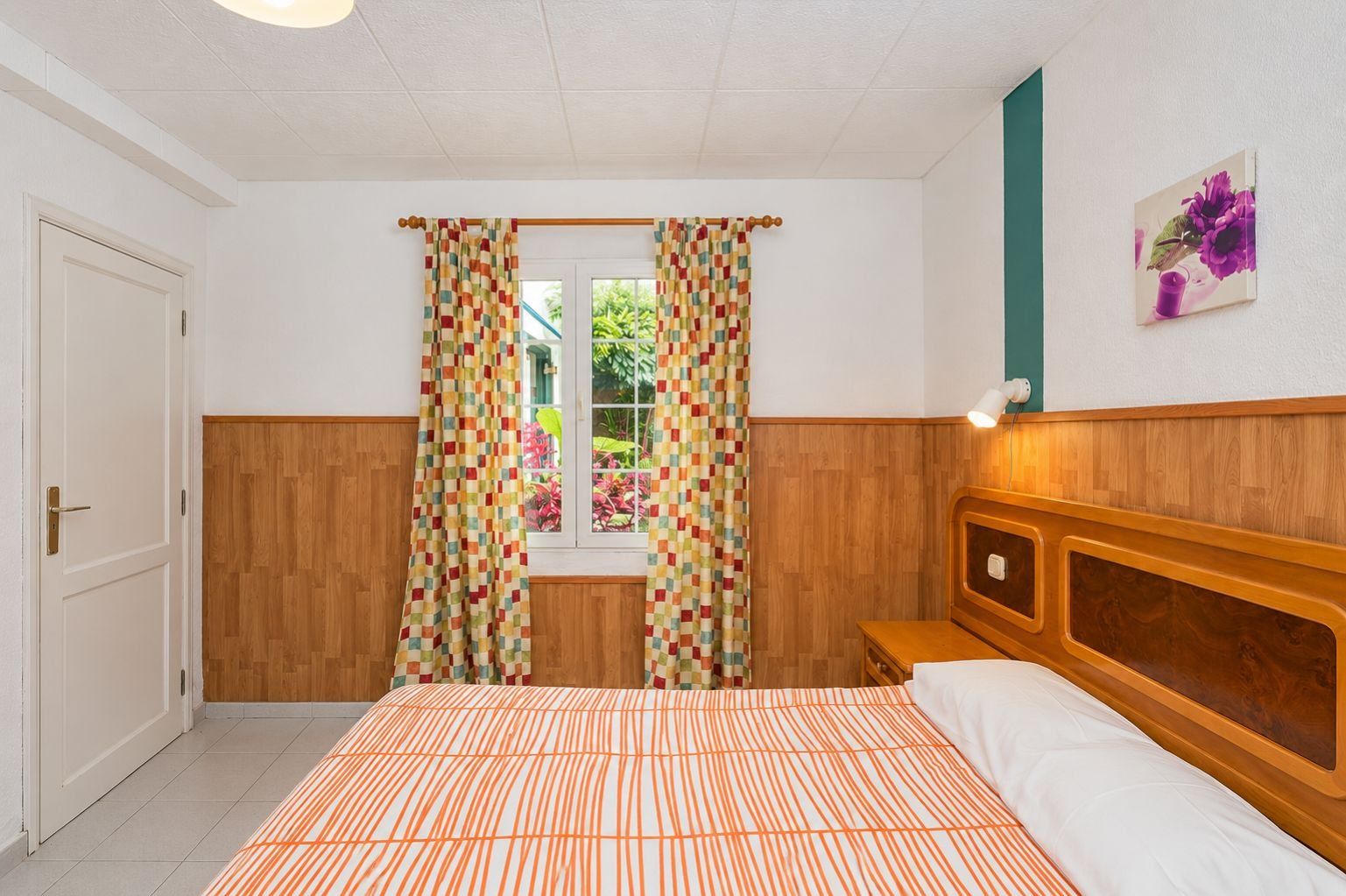 Un dormitorio con cama, cortinas, cabecero, mesita de noche, lámpara de pared y ventana con vistas a zonas verdes.