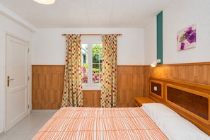 Un dormitorio con cama, cortinas, cabecero, mesita de noche, lámpara de pared y ventana con vistas a zonas verdes.
