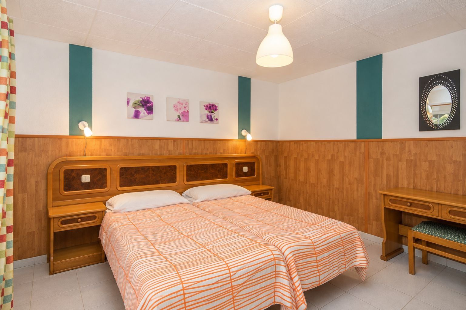 Un dormitorio con cabecera de madera, cama con colcha a rayas anaranjadas, mesita de noche, espejo y lámparas fijadas a la pared.