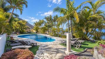 Un área de piscina con cubierta de piedra, sillas de descanso y plantas tropicales.