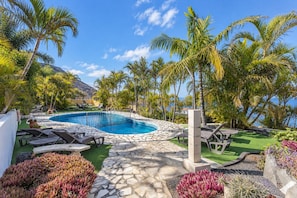 Un área de piscina con cubierta de piedra, sillas de descanso y plantas tropicales.