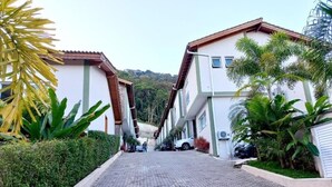 A paved pathway leading to a residential area with two-story buildings, surrounded by greenery and a hilly backdrop. - House with 3 suites on Juquehy beach. (São Sebastião)
