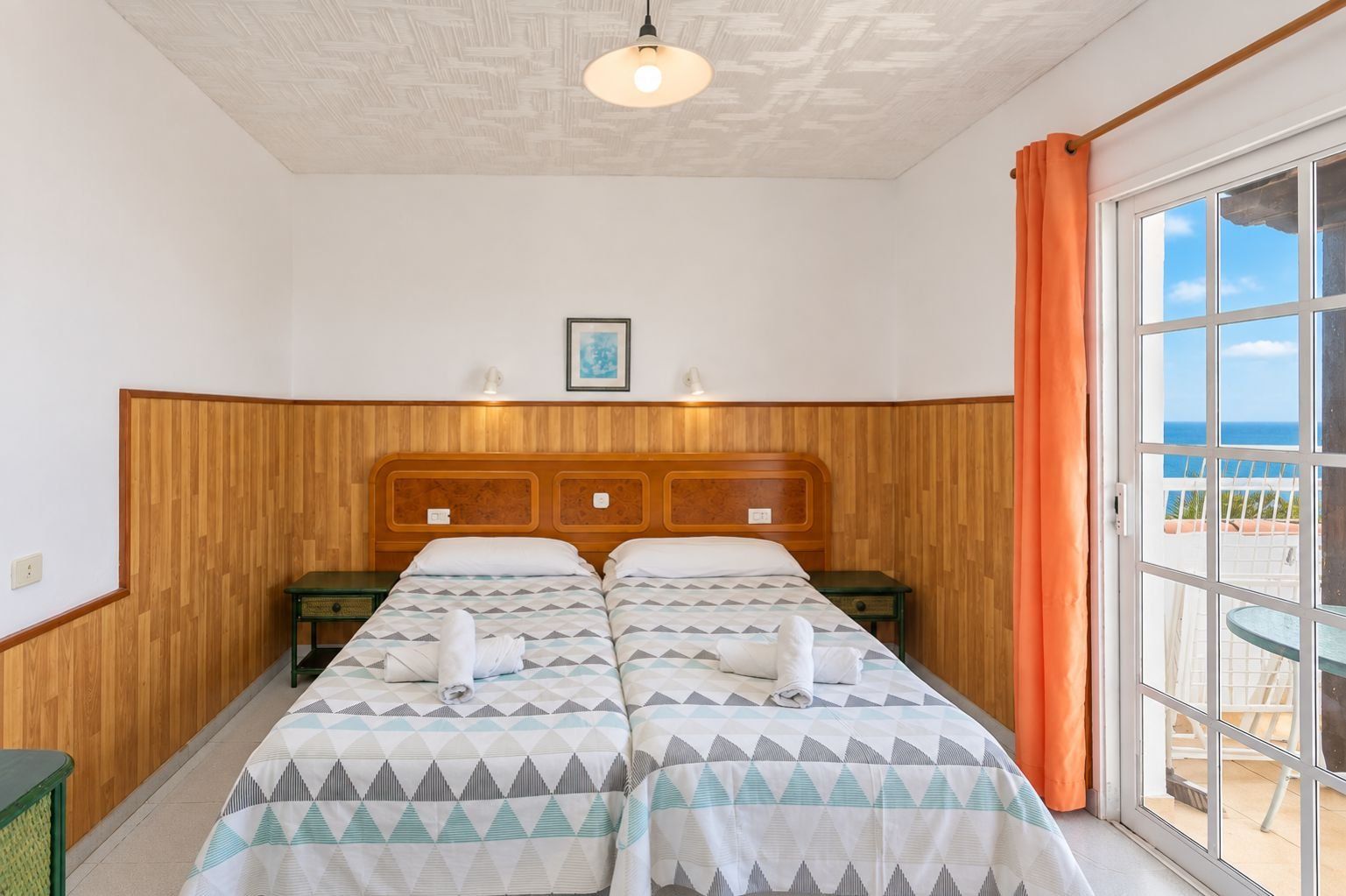 Habitación de hotel con cabecera de madera, dos camas con ropa de cama de estampado geométrico, un cuadro en la pared y vista al mar por la ventana.