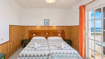 Habitación de hotel con cabecera de madera, dos camas con ropa de cama de estampado geométrico, un cuadro en la pared y vista al mar por la ventana.