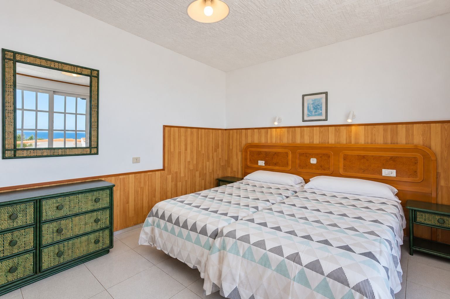 Un dormitorio con cabecera de madera, cama con ropa de cama estampada, cómoda con cajones, espejo y un cuadro en la pared.