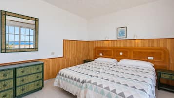 Un dormitorio con cabecera de madera, cama con ropa de cama estampada, cómoda con cajones, espejo y un cuadro en la pared.
