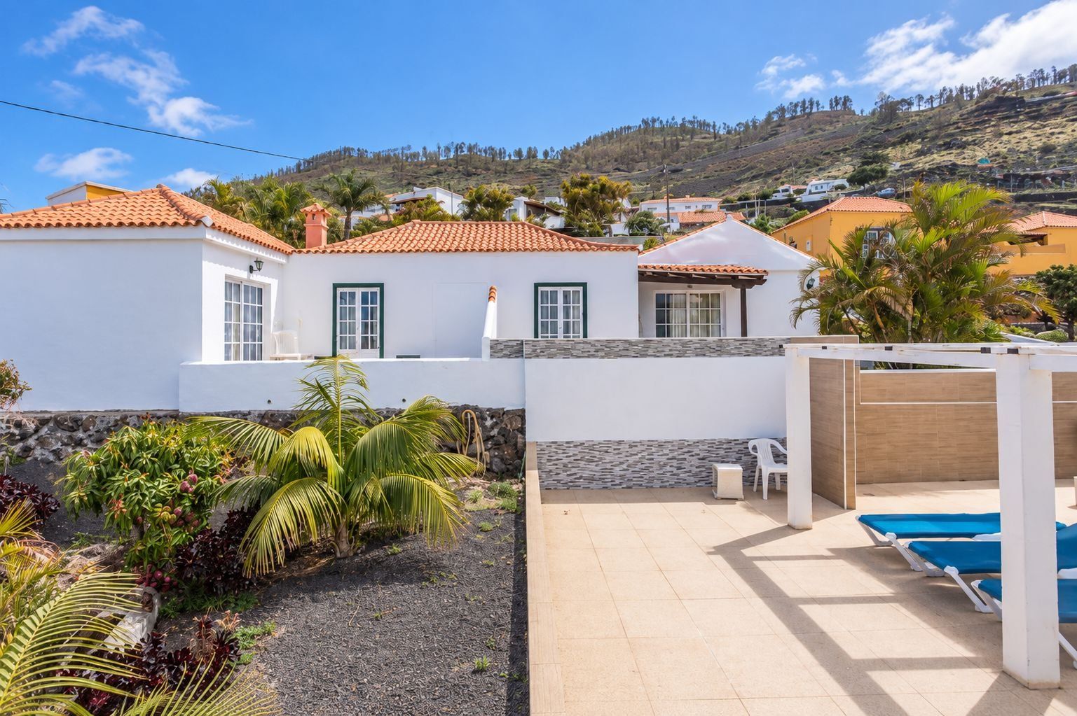 Una propiedad residencial con piscina, jardín con plantas tropicales y vistas a una colina.