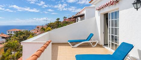 Ein Balkon mit zwei Liegestühlen, Fliesenboden und Blick auf das Meer sowie angrenzende Häuser.