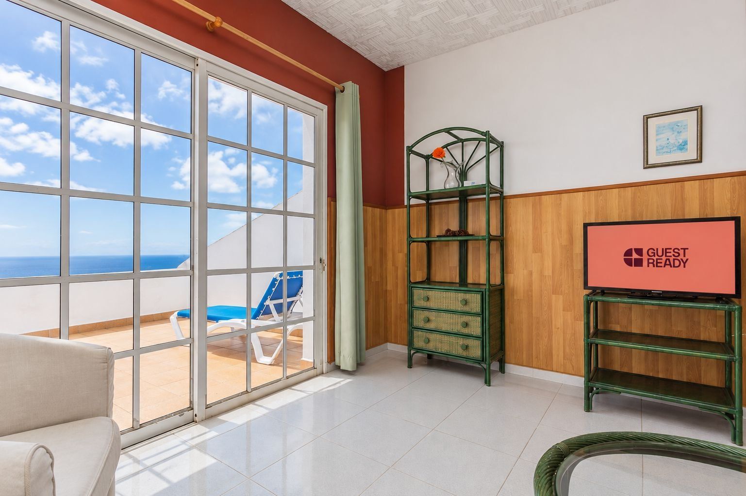 Ein Hotelzimmer mit einer Schiebetür zu einem Balkon, ausgestattet mit einem blauen Stuhl, einem grünen Korbtisch und einem Fernseher auf einem Schrank. Auf dem Schrank steht 'GUEST READY'.