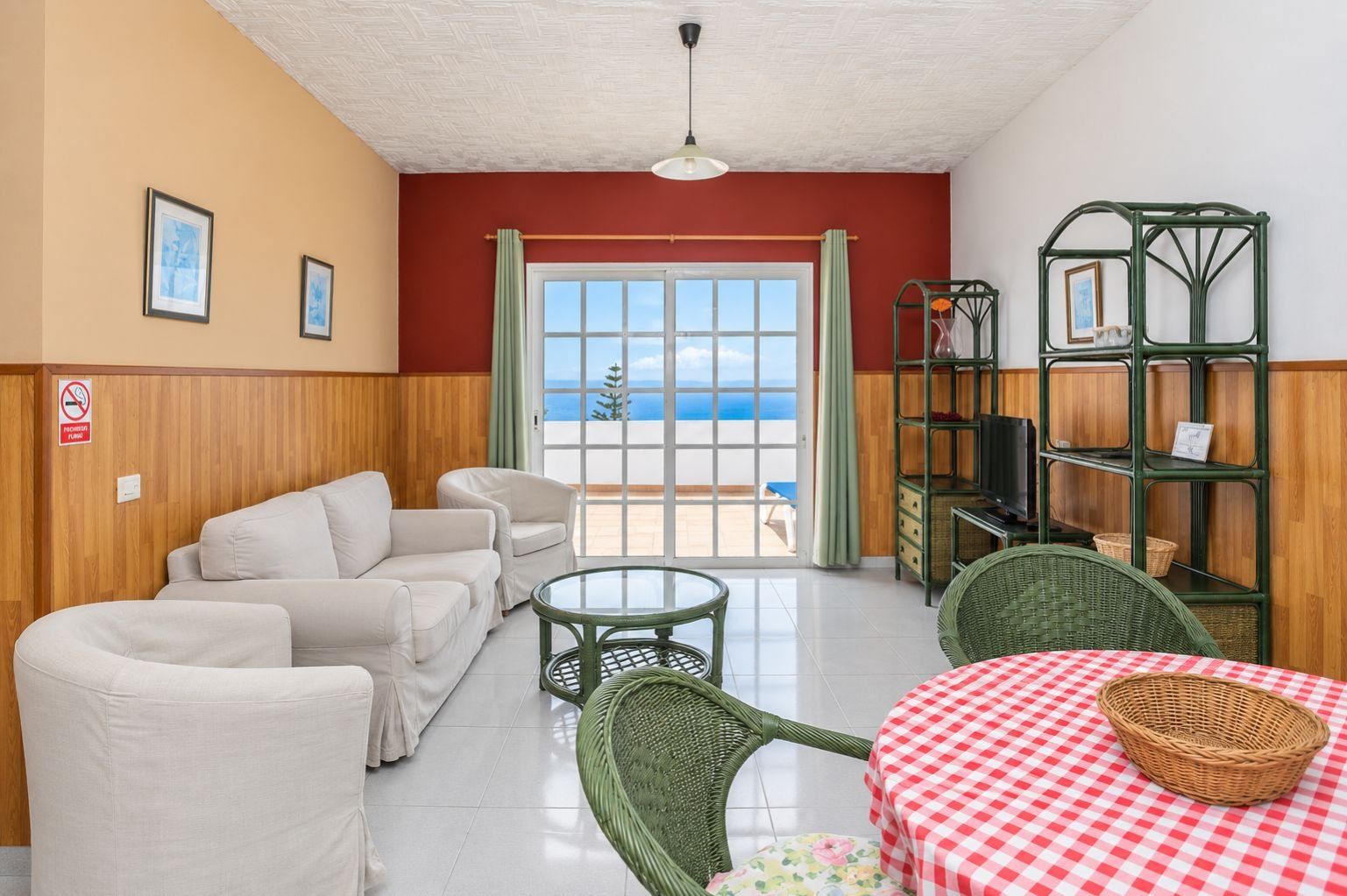 A room with a sofa set, a table with a checkered tablecloth, wicker chairs, and a green metal shelf unit.