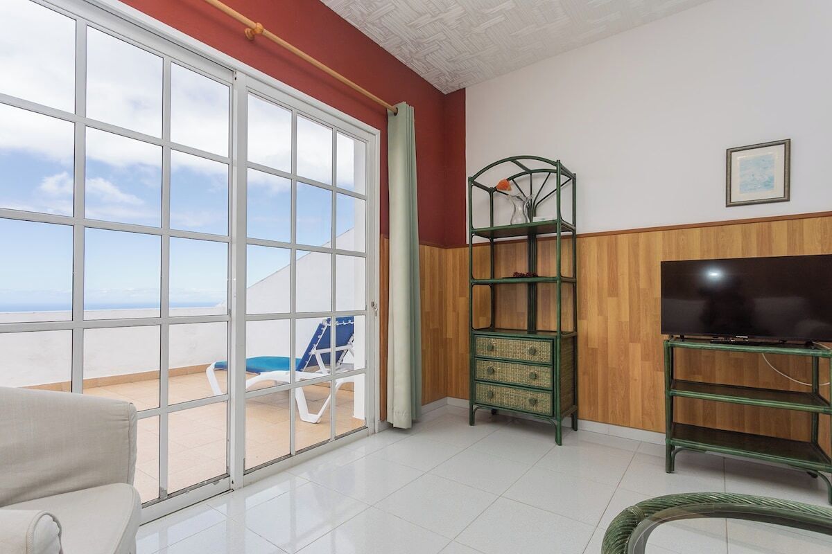 Een kamer met een grote schuifpui die uitkomt op een balkon, een televisie op een houten standaard en een rieten stoel op het balkon.