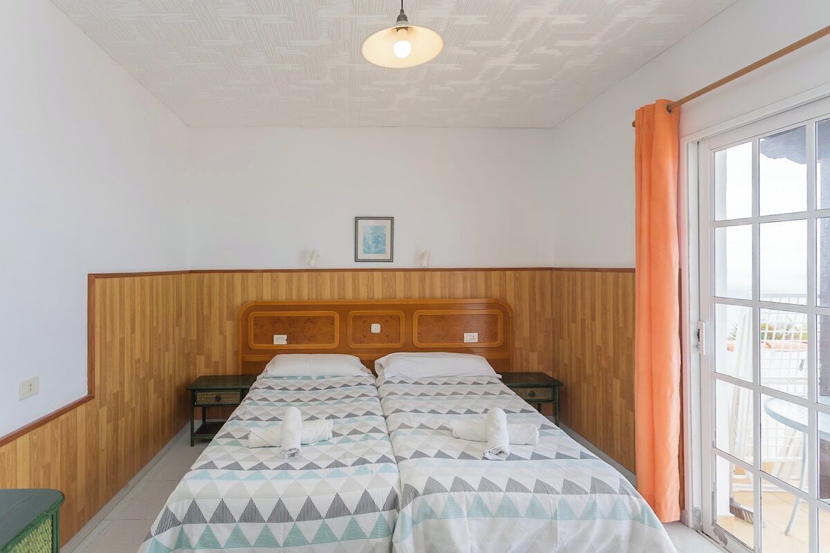 Een hotelkamer met een houten hoofdbord, twee bedden met beddengoed met een patroon, een klein nachtkastje en een groot raam met gordijnen.