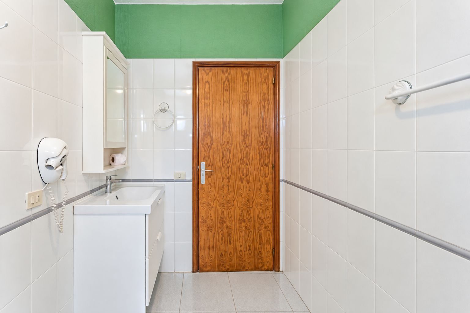 A bathroom with white tiles, a wooden door, a white sink, a mirror, and a towel dispenser.