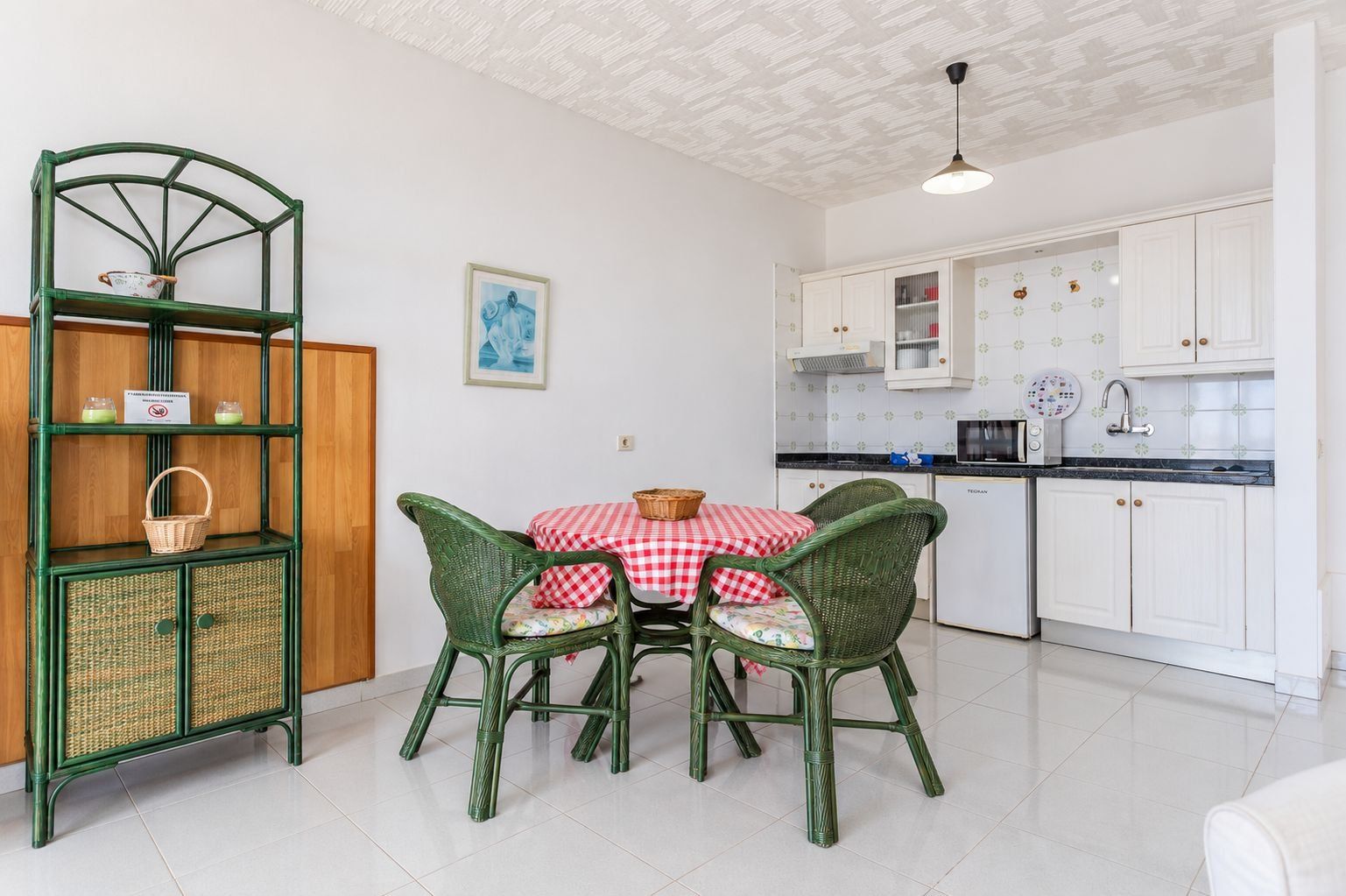 Cocina compacta con armarios blancos, comedor con sillas de mimbre verde y mantel a cuadros rojos y blancos.