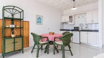Cocina compacta con armarios blancos, comedor con sillas de mimbre verde y mantel a cuadros rojos y blancos.