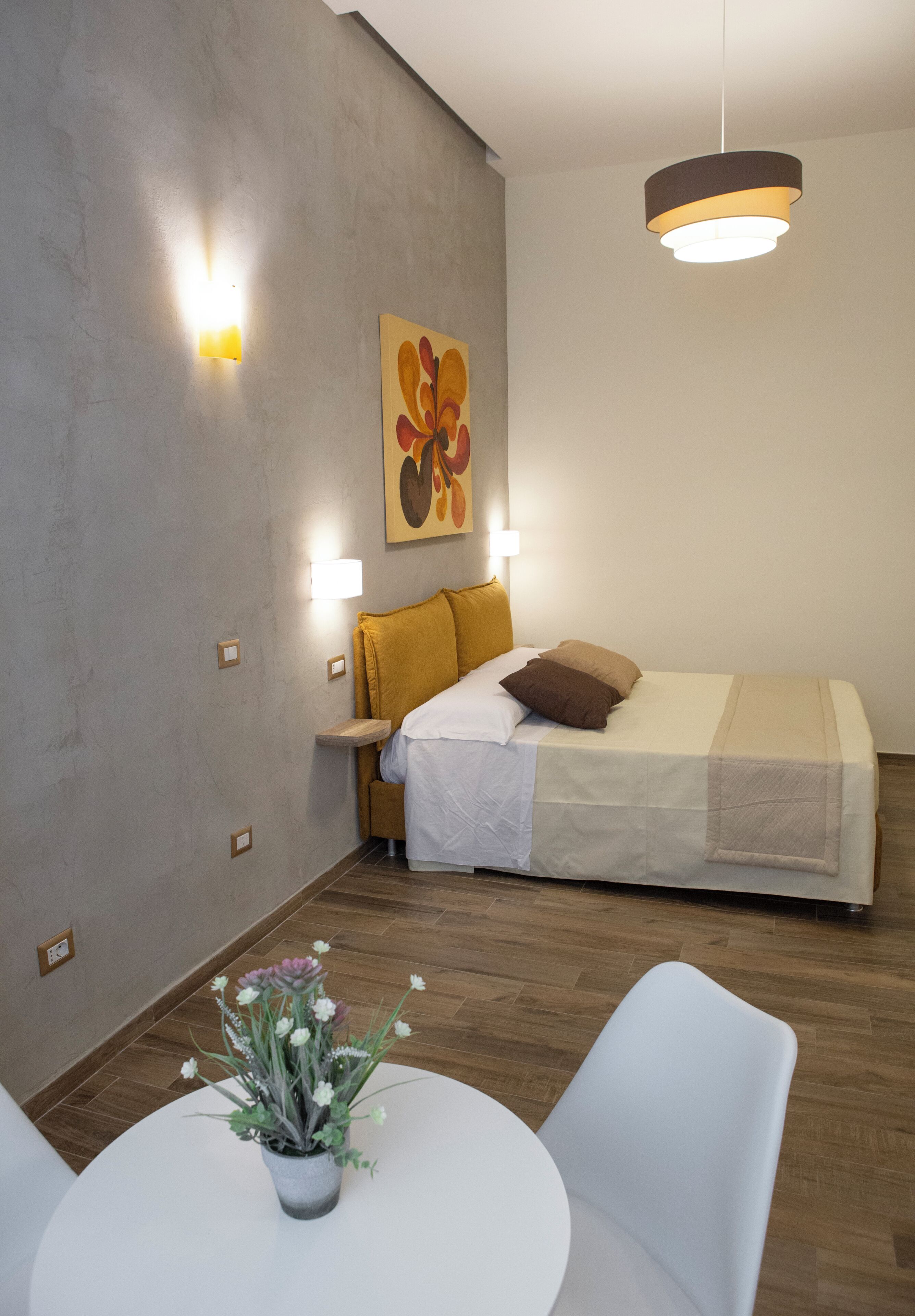 A modern bedroom with a bed, bedside tables, a wall-mounted artwork, a hanging light fixture, and a round dining table with chairs.