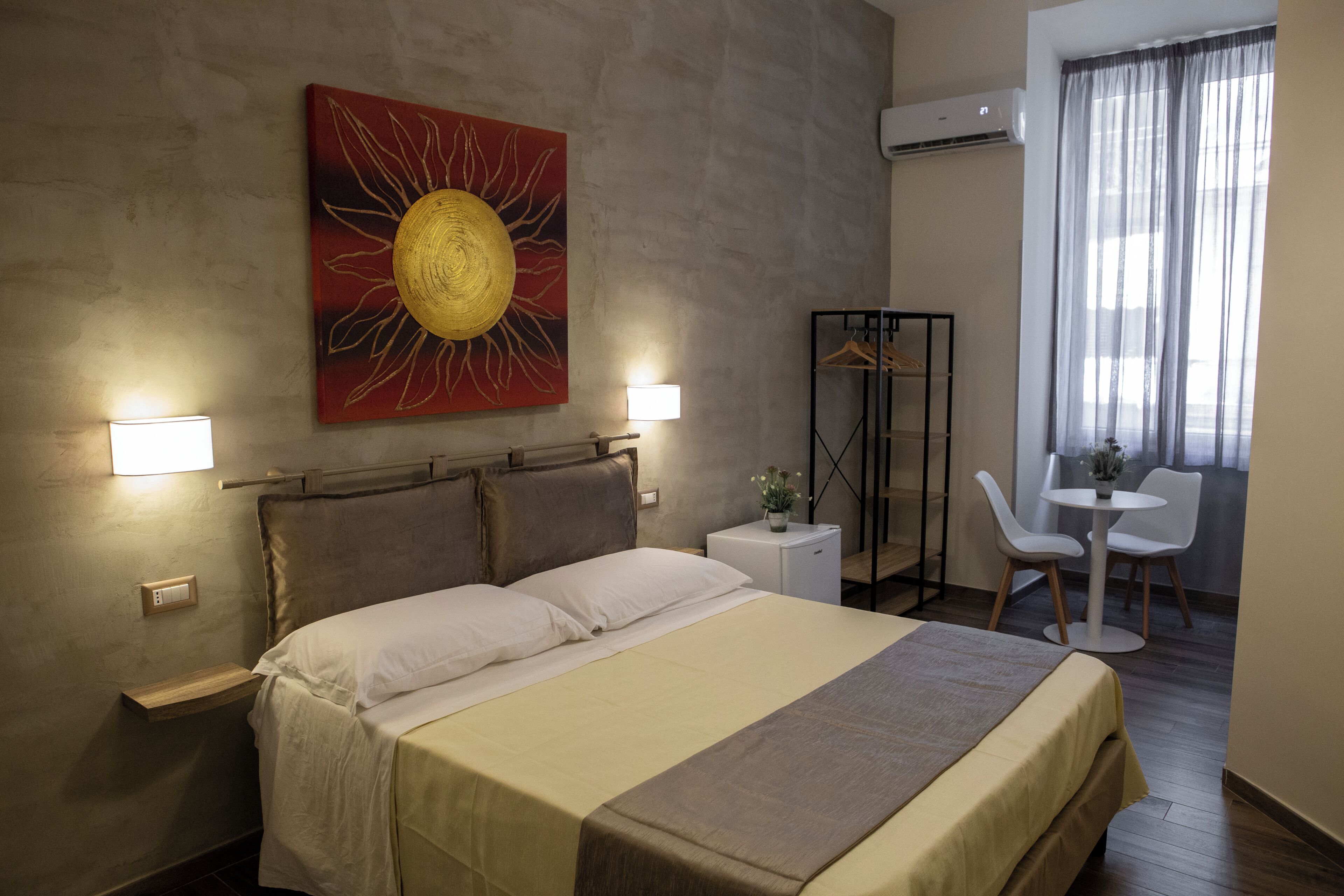 A hotel room with a bed, bedside lamps, a small table with a chair, an air conditioning unit, and a window with curtains.