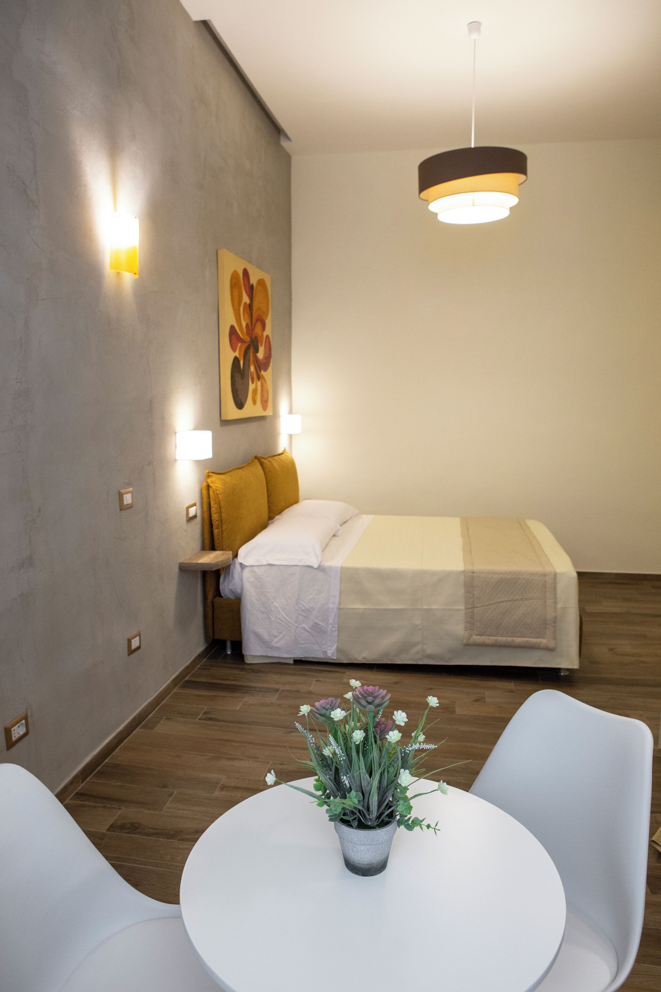 A hotel room with a bed, bedside lamps, a painting, a round table with chairs, and a potted plant.