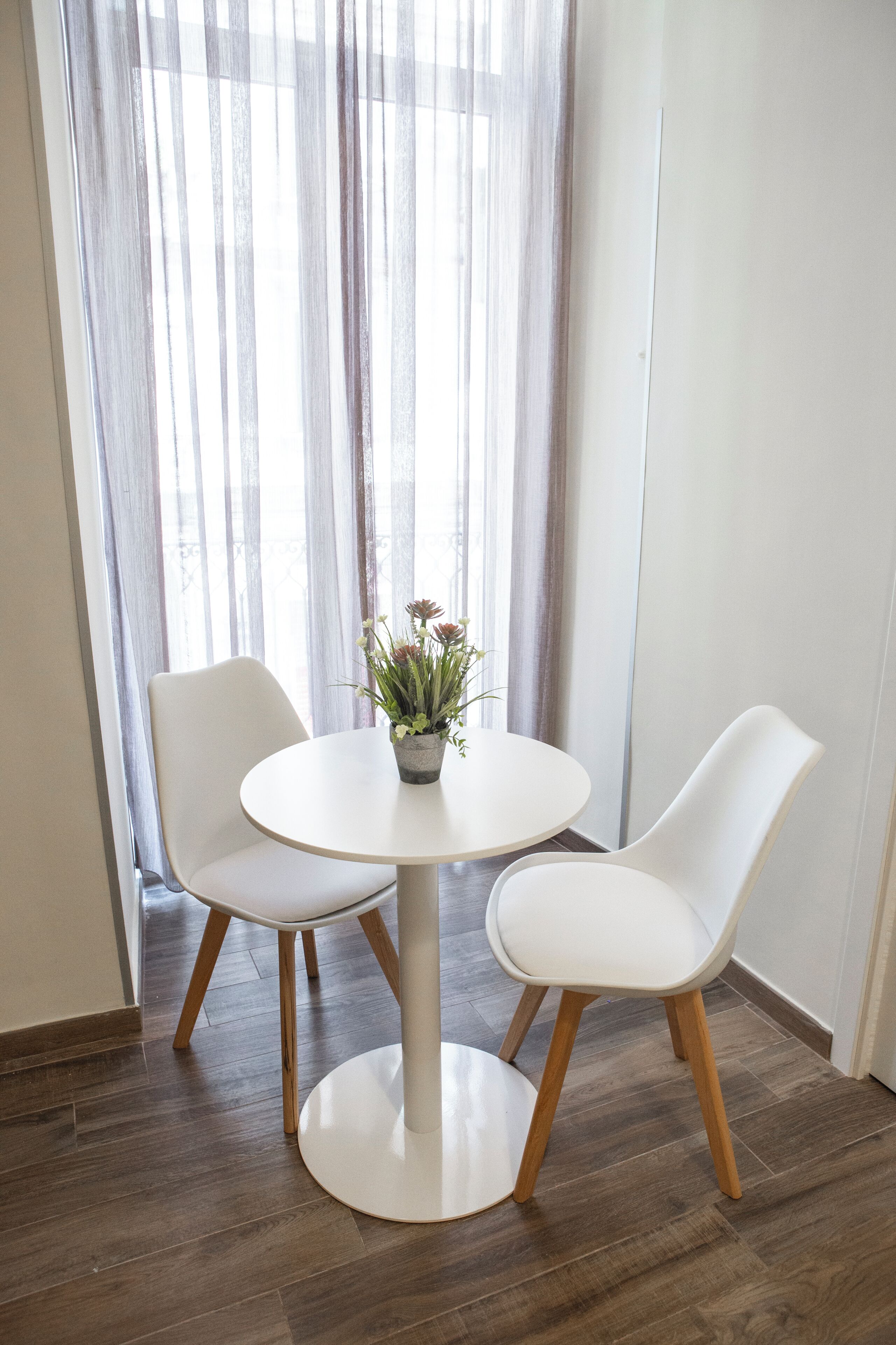 Une table ronde, deux chaises blanches, une plante en pot et une fenêtre ornée de rideaux légers.