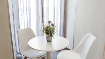 Une table ronde, deux chaises blanches, une plante en pot et une fenêtre ornée de rideaux légers.