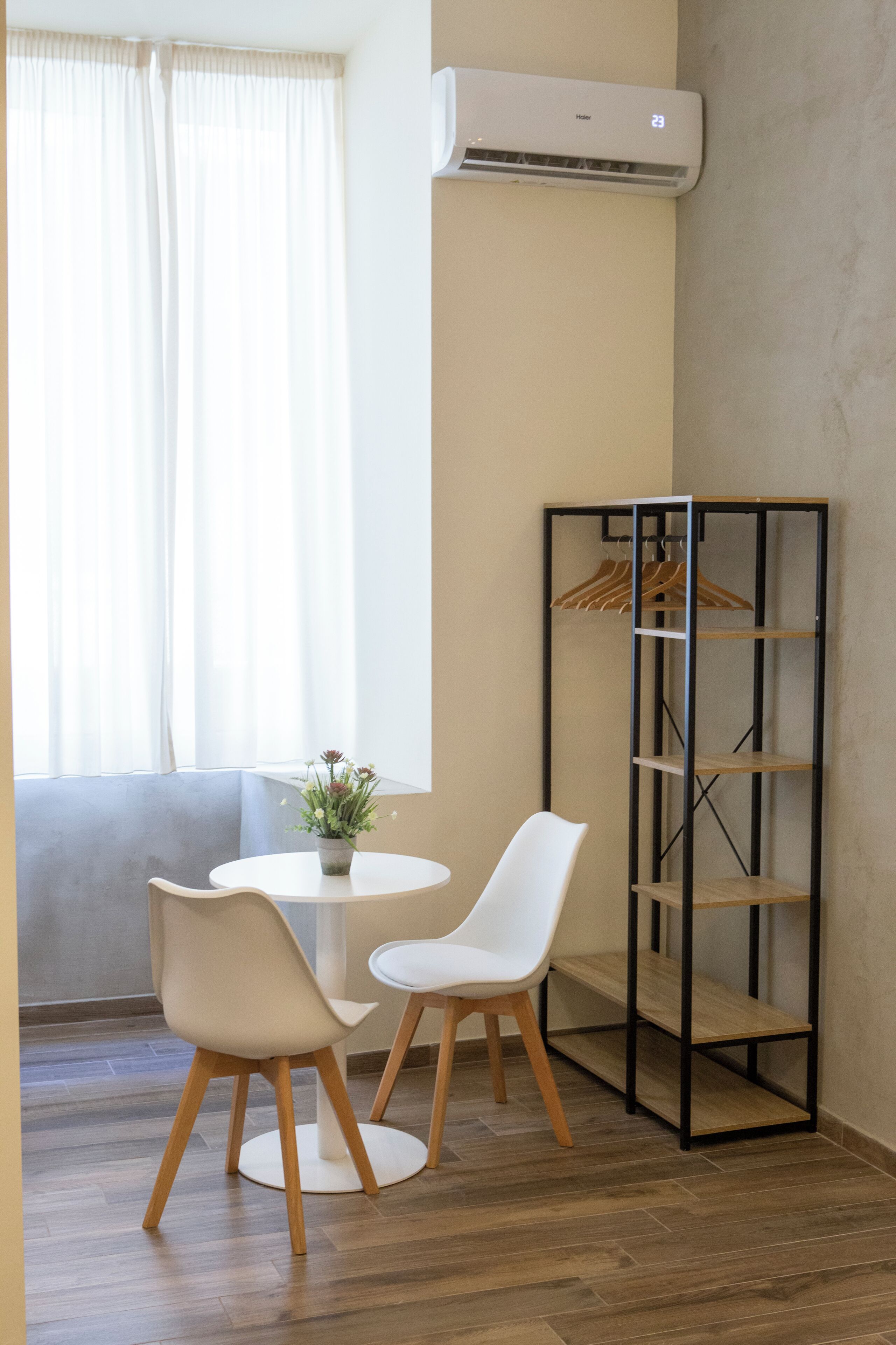 Une pièce comprenant une table ronde, deux chaises, une plante en pot, un climatiseur fixé au mur et une étagère métallique où sont rangés des vêtements.