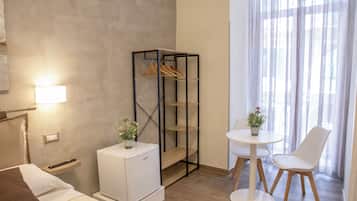 Une chambre d’hôtel avec un lit, une petite table blanche et deux chaises, un mini-réfrigérateur, un porte-manteau et une fenêtre avec des rideaux légers.