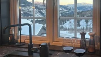 Eine Küche mit Blick auf schneebedeckte Berge, ein Spülbecken, ein Teller mit Käse und Wurstwaren sowie zwei Gläsern Rotwein.