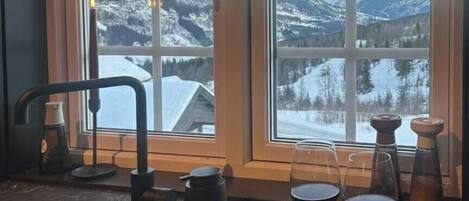 Une cuisine donnant sur des montagnes enneigées, un évier, une assiette de fromages et de charcuteries, et deux verres de vin rouge.