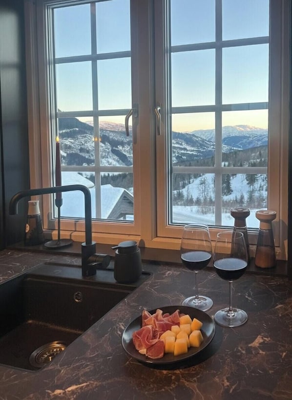 Una cocina con vistas a montañas nevadas, un lavamanos, un plato de queso y embutidos, y dos copas de vino tinto.