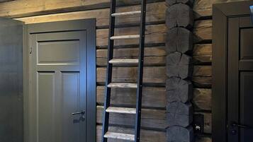 Un intérieur en bois avec une échelle métallique adossée à un mur de pierre, deux portes sombres et un plafond en pente.
