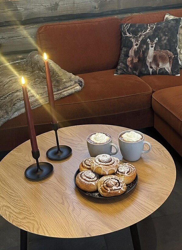 Un coin salon accueillant avec une table ronde en bois, deux bougies, une assiette de pâtisseries et deux tasses de café.