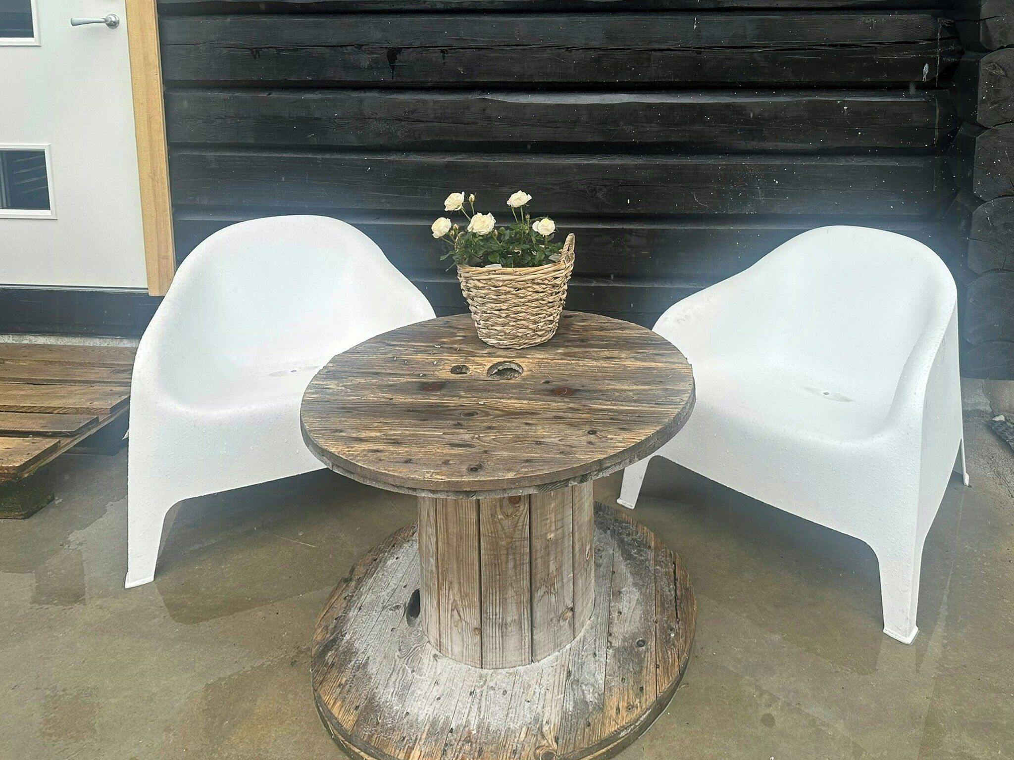 A round wooden table with a woven basket containing white flowers, flanked by two white plastic chairs.