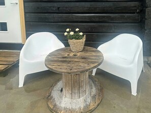 Une table ronde en bois sur laquelle repose un panier tressé rempli de fleurs blanches, et deux chaises en plastique blanc placées de part et d’autre.