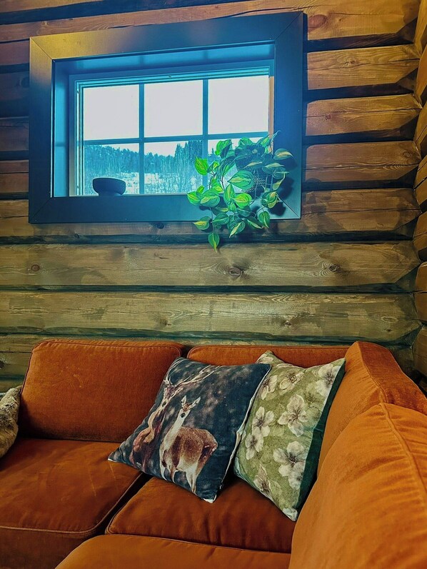 A log cabin interior with a window overlooking a snowy landscape, a cushioned sofa with decorative pillows, and a potted plant on the windowsill.
