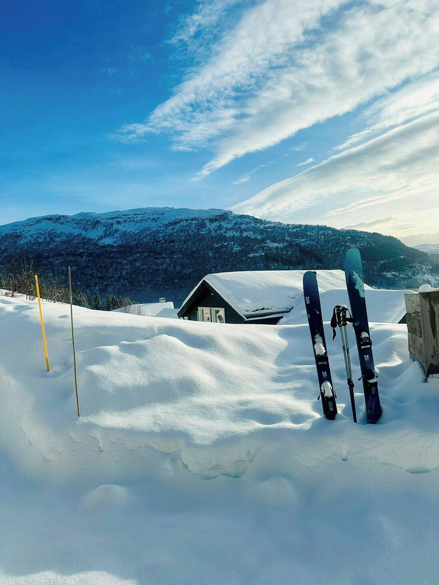 Paesaggio innevato con una baita di legno e due paia di sci appoggiati a un palo di legno.