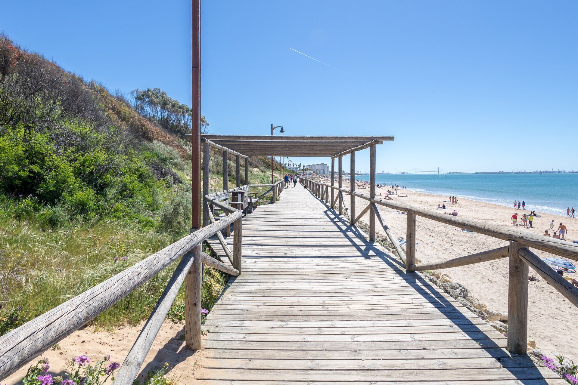 Un paseo marítimo de madera que lleva a una playa con gente y un cielo despejado.