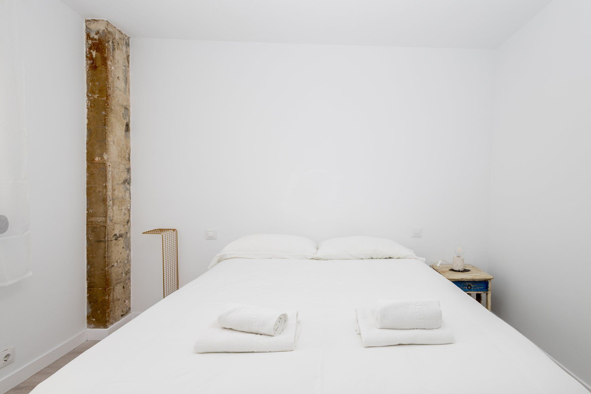 Ein schlicht eingerichtetes Schlafzimmer mit einem Einzelbett, zwei zusammengerollten Handtüchern, einem Nachttisch und einer Wandlampe.