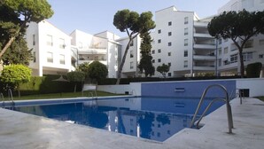 Una piscina con una escalera de metal, rodeada por un solárium de hormigón, con edificios al fondo.