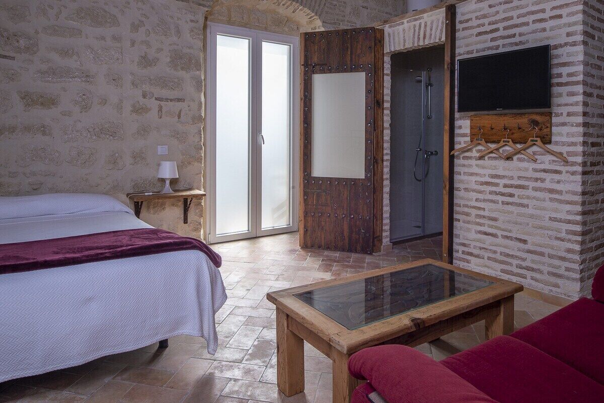 A room with a bed, a glass-top table, a red sofa, a wooden door, and a television mounted on a brick wall.